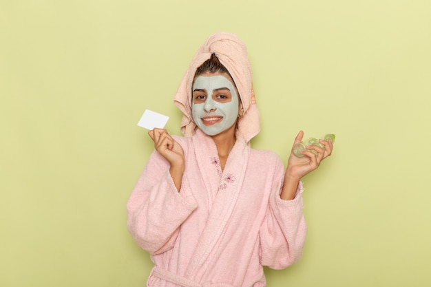Mulher jovem de frente após o banho com roupão rosa segurando sprays e cartão na mesa verde