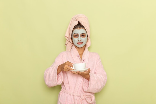 Mulher jovem de frente após o banho com roupão rosa tomando café na mesa verde