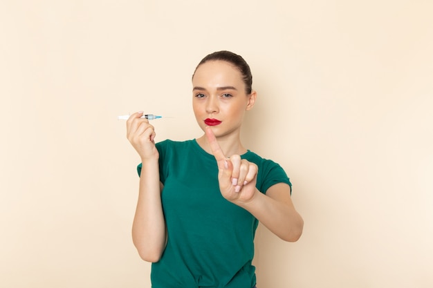 Mulher jovem de frente com camisa verde escura e calça jeans segurando a injeção na mesa bege.