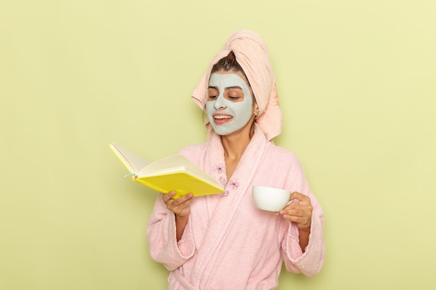 Mulher jovem, de frente, depois do banho, com um roupão rosa, bebendo café e lendo o caderno na superfície verde