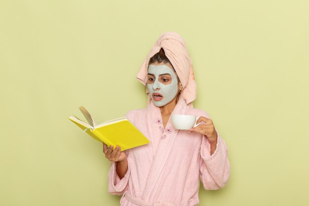 Mulher jovem, de frente, depois do banho, com um roupão rosa, bebendo café e lendo o caderno na superfície verde