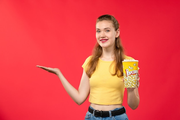 Foto grátis mulher jovem de frente no cinema segurando pipoca e sorrindo na parede vermelha.