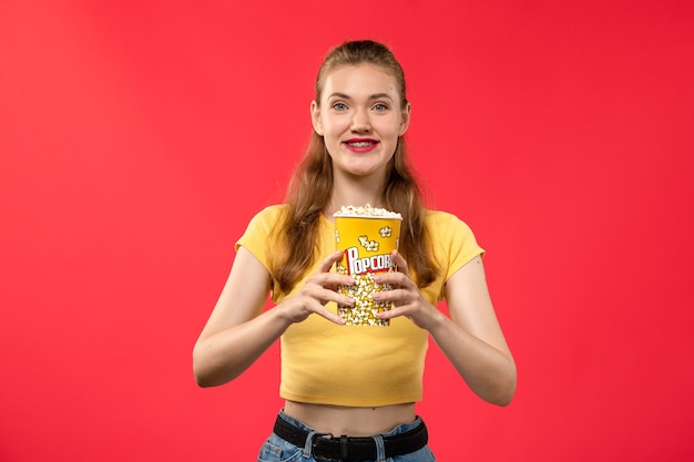 Mulher jovem de frente no cinema segurando um pacote de pipoca e sorrindo na parede vermelha filmes teatro cinema lanche feminino divertido filme