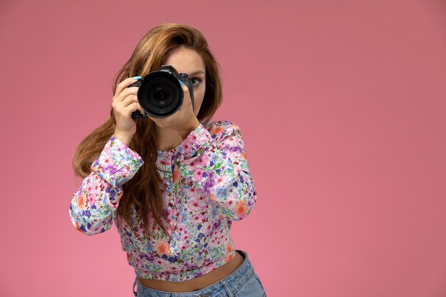Mulher jovem de frente para a camisa com design flor e calça jeans, sorrindo, tirando uma foto com a câmera no fundo rosa