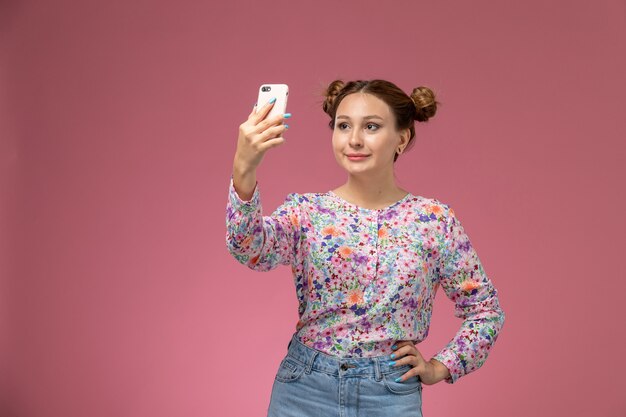 Mulher jovem de frente para a camisa com design flor e calça jeans, tirando selfie no fundo rosa
