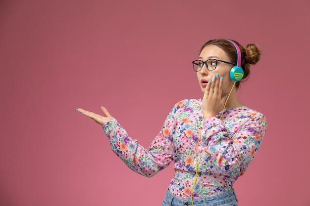 Mulher jovem de frente para a camisa com design floral e jeans azul, ouvindo música com fones de ouvido no fundo rosa, pose feminina