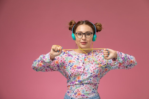 Mulher jovem de frente para a camisa com design floral e jeans azul, ouvindo música com fones de ouvido no fundo rosa