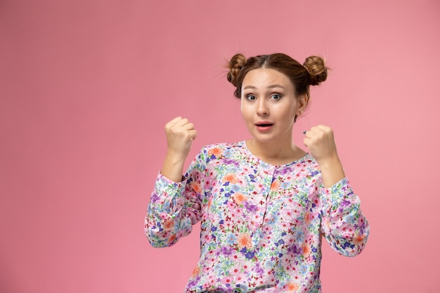 Mulher jovem de frente para a camisa com design floral e jeans, posando com uma expressão encantada no fundo rosa