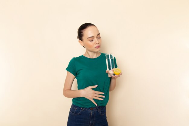 Mulher jovem de frente para a camisa verde escura e calça jeans azul segurando um copo de água na cor bege