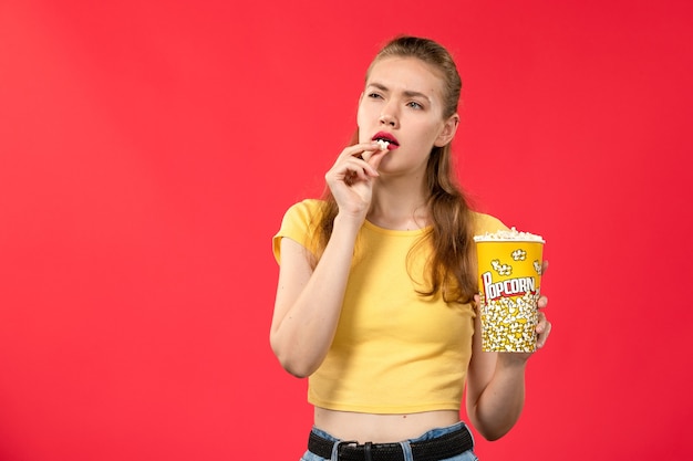 Mulher jovem de frente para o cinema segurando e comendo pacote de pipoca no filme de parede vermelha-clara.