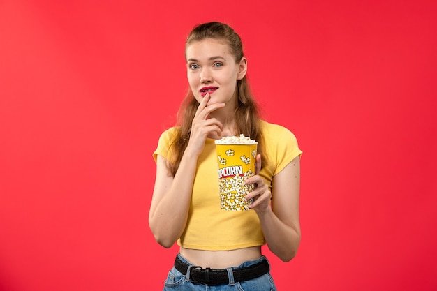 Mulher jovem de frente para o cinema segurando pipoca na parede vermelha.
