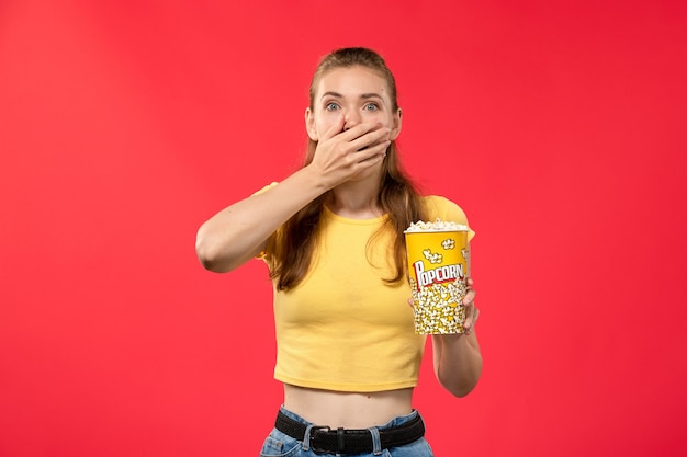 Foto grátis mulher jovem de frente para o cinema segurando um pacote de pipoca com expressão de choque na parede vermelha filmes teatro cinema lanche divertido filme