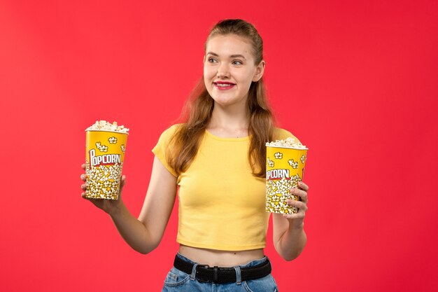 Mulher jovem de frente para o cinema segurando um pacote de pipoca com um sorriso na parede vermelha-clara.