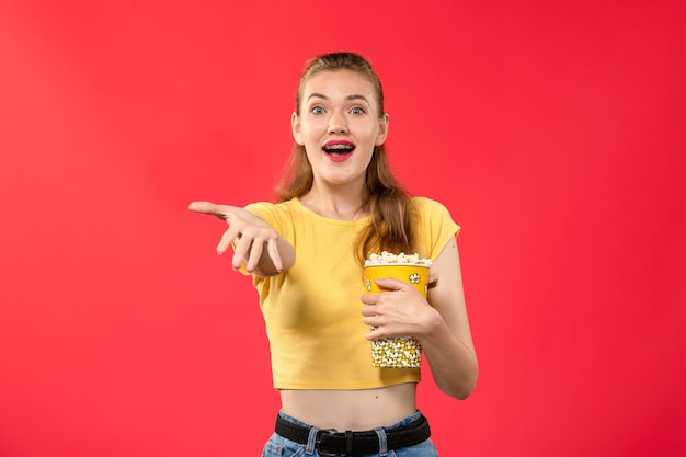 Mulher jovem de frente para o cinema segurando um pacote de pipoca na parede vermelho-claro.