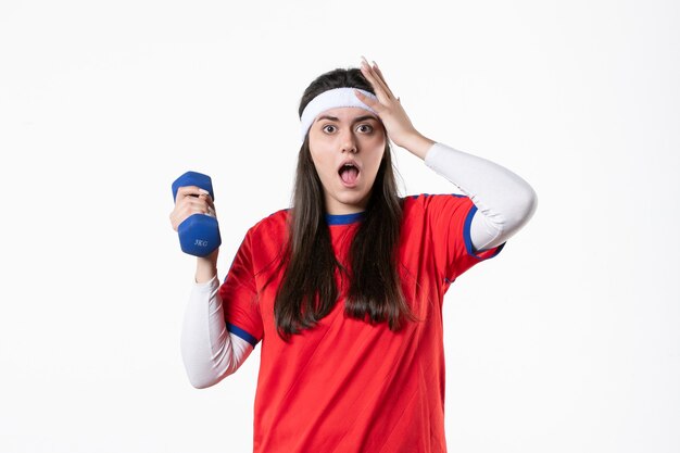 Mulher jovem de frente para o esporte com roupas esportivas, malhando com halteres na parede branca