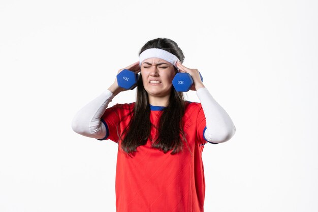 Mulher jovem de frente para o esporte com roupas esportivas, malhando com halteres na parede branca