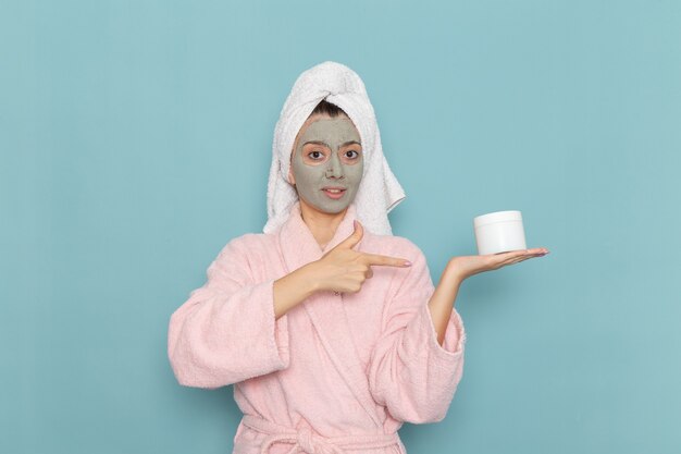 Foto grátis mulher jovem de frente para um roupão rosa com máscara no rosto, segurando o creme na parede azul claro, chuveiro, creme de limpeza de beleza para cuidados pessoais