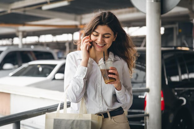 Mulher jovem de negócios com café falando alegremente ao telefone no estacionamento