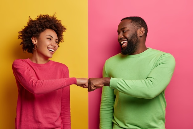 Mulher jovem de pele escura positiva e homem batem os punhos, concordam em ser uma equipe, olham felizes um para o outro, comemora a tarefa concluída, usa roupas rosa e verdes, posa em ambientes fechados, tem um negócio bem-sucedido