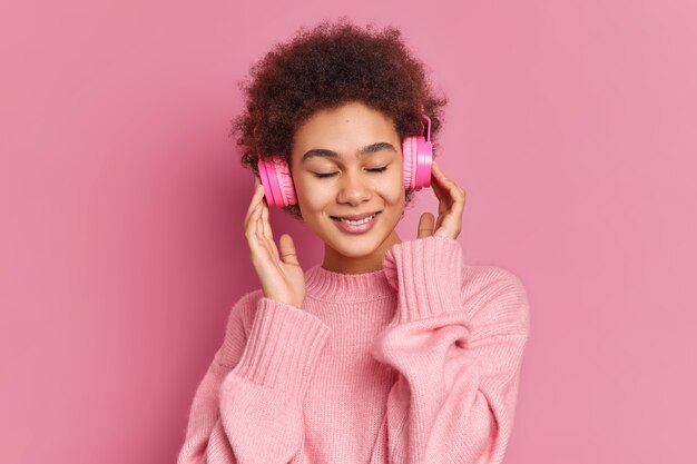 Mulher jovem de pele escura satisfeita gosta de ouvir uma melodia agradável mantém as mãos nos fones de ouvido estéreo fecha os olhos e usa um macacão casual