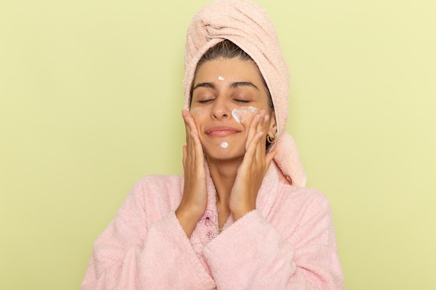 Mulher jovem de roupão rosa aplicando creme facial em uma superfície verde clara