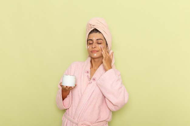 Mulher jovem de roupão rosa aplicando creme facial na superfície verde