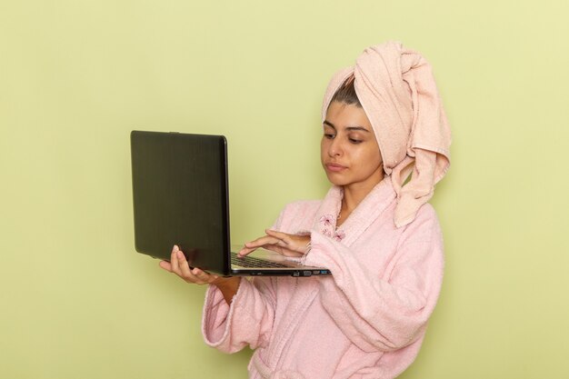 Mulher jovem de roupão rosa, de frente, usando laptop na superfície verde