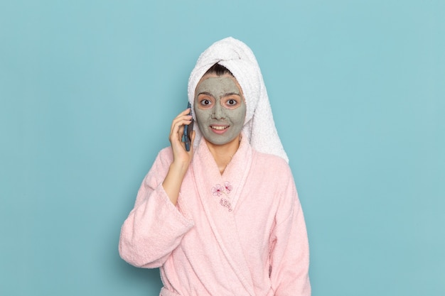 Mulher jovem de roupão rosa depois do banho, de frente, falando ao telefone na parede azul.