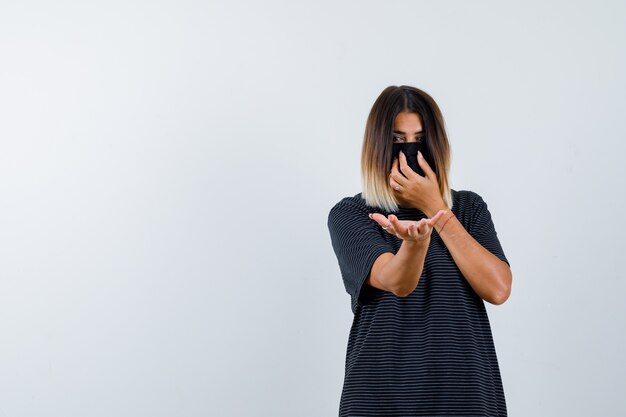 Mulher jovem de vestido preto, máscara preta esticando uma mão em direção à câmera segurando algo, colocando a outra mão na máscara e olhando séria, vista frontal.