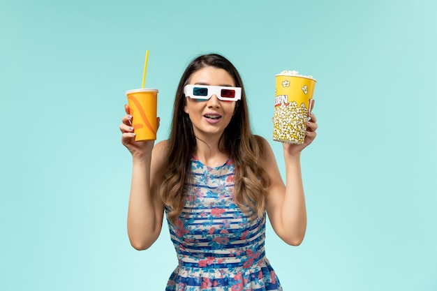 Mulher jovem de vista frontal com pacote de pipoca e bebida em óculos de sol d na superfície azul