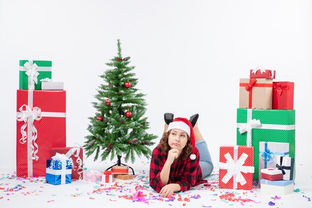 Mulher jovem deitada de frente para os presentes de Natal e uma pequena árvore de férias no fundo branco ano novo frio mulher natal neve