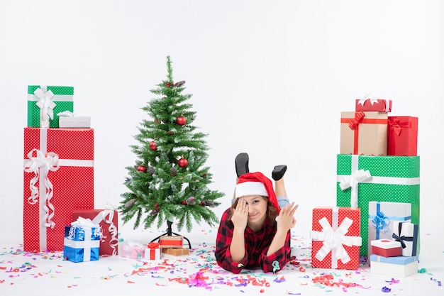 Mulher jovem deitada de frente para os presentes de natal e uma pequena árvore de férias no fundo branco.