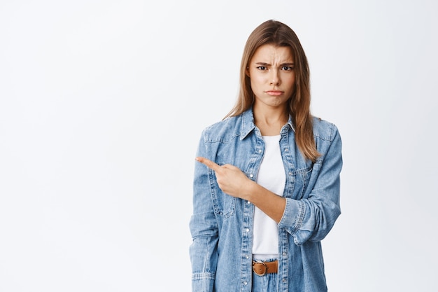 Mulher jovem descontente e chateada, carrancuda, ofendida e reclamando, apontando o dedo para a esquerda para o espaço vazio em branco, em pé contra a parede do estúdio