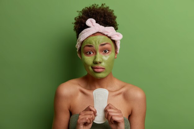 Mulher jovem descontente segura absorvente higiênico para a menstruação, aplica máscara facial de beleza para rejuvenescimento, usa bandana e toalha, posa dentro de casa contra uma parede verde. Mulheres, beleza, conceito de higiene