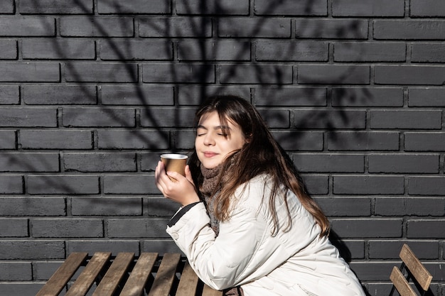 Mulher jovem, desfrutando de uma bebida contra a parede de tijolos do exterior da cafeteria.