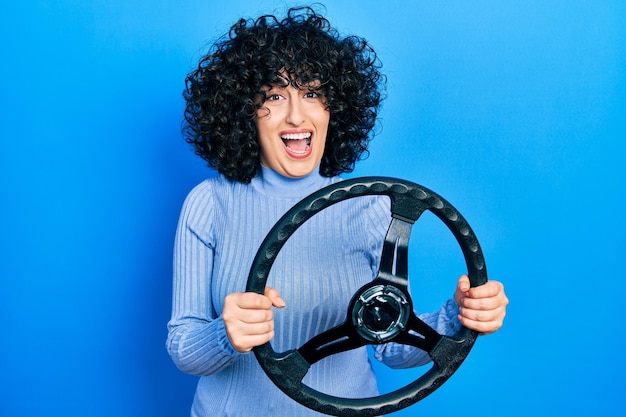 Mulher jovem do Oriente Médio segurando o volante comemorando louco e espantado pelo sucesso com os olhos abertos gritando animado.