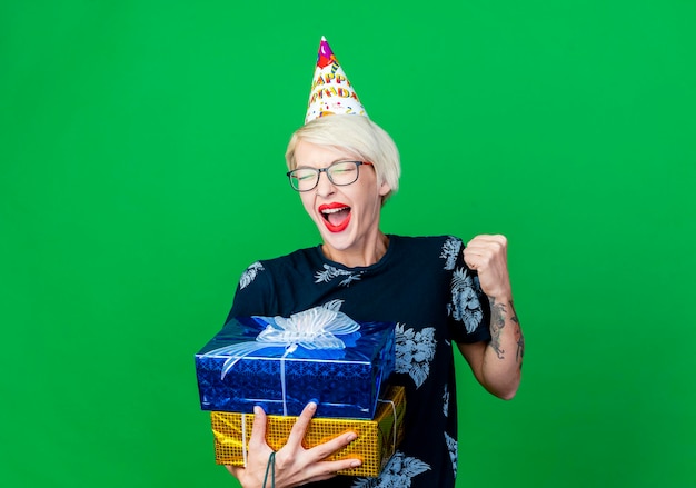 Mulher jovem e alegre de festa loira usando óculos e boné de aniversário segurando caixas de presente, fazendo gesto de sim, isolado na parede verde com espaço de cópia