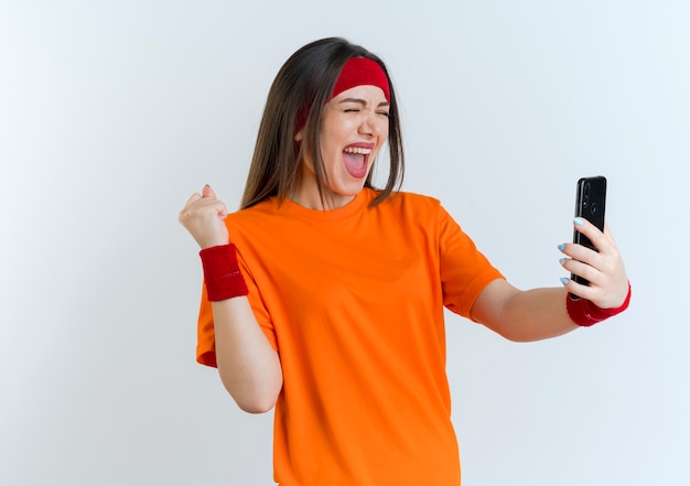 Mulher jovem e alegre e esportiva usando bandana e pulseiras segurando um telefone celular, fazendo gesto de sim com os olhos fechados, isolado na parede branca com espaço de cópia