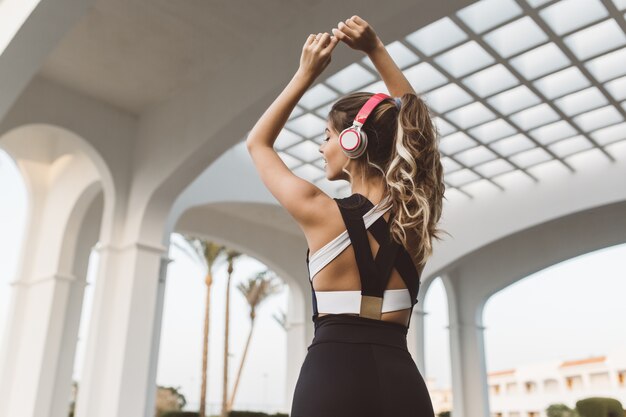 Mulher jovem e alegre incrível no sportswear das costas, esticando as mãos acima do lado de fora na arquitetura branca do leste. Expressar positividade, estilo de vida ativo, praticar esportes, ouvir música.