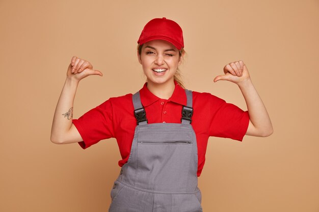 Mulher jovem e alegre, trabalhadora da construção civil, vestindo uniforme e boné apontando para si mesma, piscando