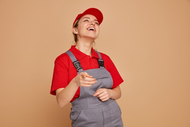 Mulher jovem e alegre trabalhadora da construção civil vestindo uniforme e boné olhando para cima apontando para a câmera rindo