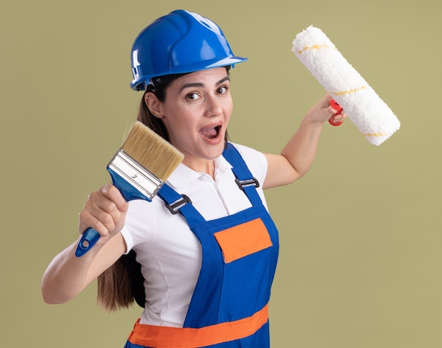 Mulher jovem e animada construtora de uniforme segurando a escova giratória e o pincel isolado na parede verde oliva