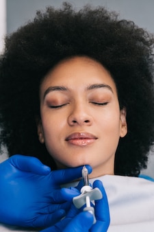 Mulher jovem e atraente africana está recebendo injeções faciais rejuvenescedoras. ela está calmamente sentada na clínica. a esteticista especialista está preenchendo as rugas femininas com ácido hialurônico.