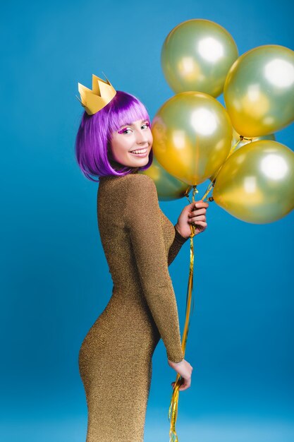 Mulher jovem e atraente alegre num vestido elegante de luxo, celebrando a grande festa. Balões dourados, coroa, corte de cabelo roxo, maquiagem brilhante, sorrindo, feriados de celebração.