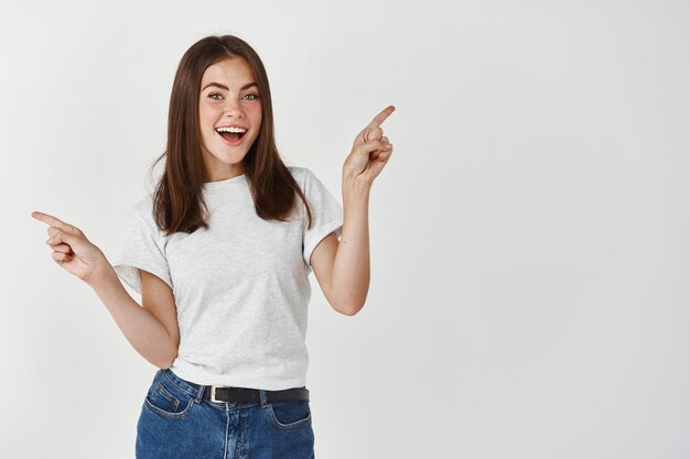 Mulher jovem e atraente apontando os dedos para o lado, mostrando duas variantes, demonstrar banners de produtos e sorrindo, em pé sobre uma parede branca.