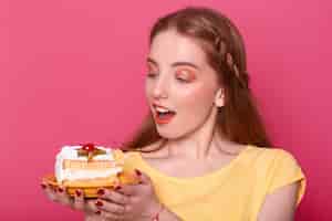 Foto grátis mulher jovem e atraente com a boca aberta mantém o prato com um pedaço de bolo delicioso nas mãos. senhora de cabelos castanho com manicure vermelho