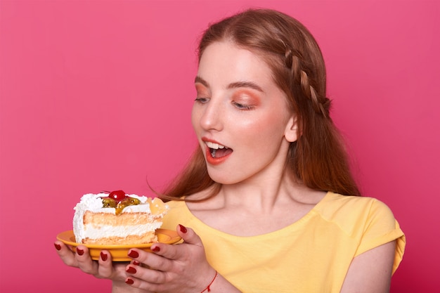 Mulher jovem e atraente com a boca aberta mantém o prato com um pedaço de bolo delicioso nas mãos. Senhora de cabelos castanho com manicure vermelho