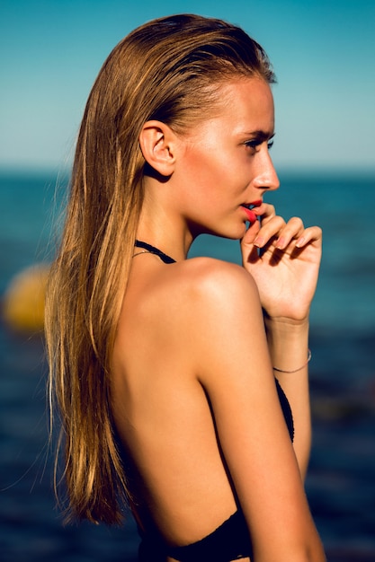 Mulher jovem e atraente com bronzeado perfeito fit corpo posando na praia com céu azul.