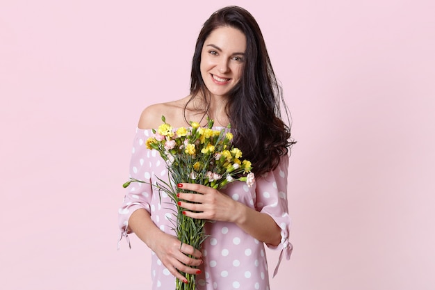 Mulher jovem e atraente com cabelo preto longo ondulado, detém flores, vestida com um vestido de bolinhas, tem clima de primavera, posa em rosa claro, tem encontro romântico com o namorado. 8 de março de conceito