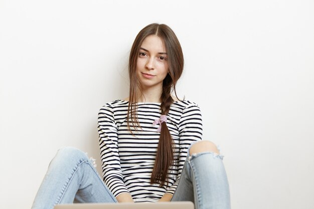 Mulher jovem e atraente com camiseta listrada de mangas compridas e jeans rasgados sentada no chão